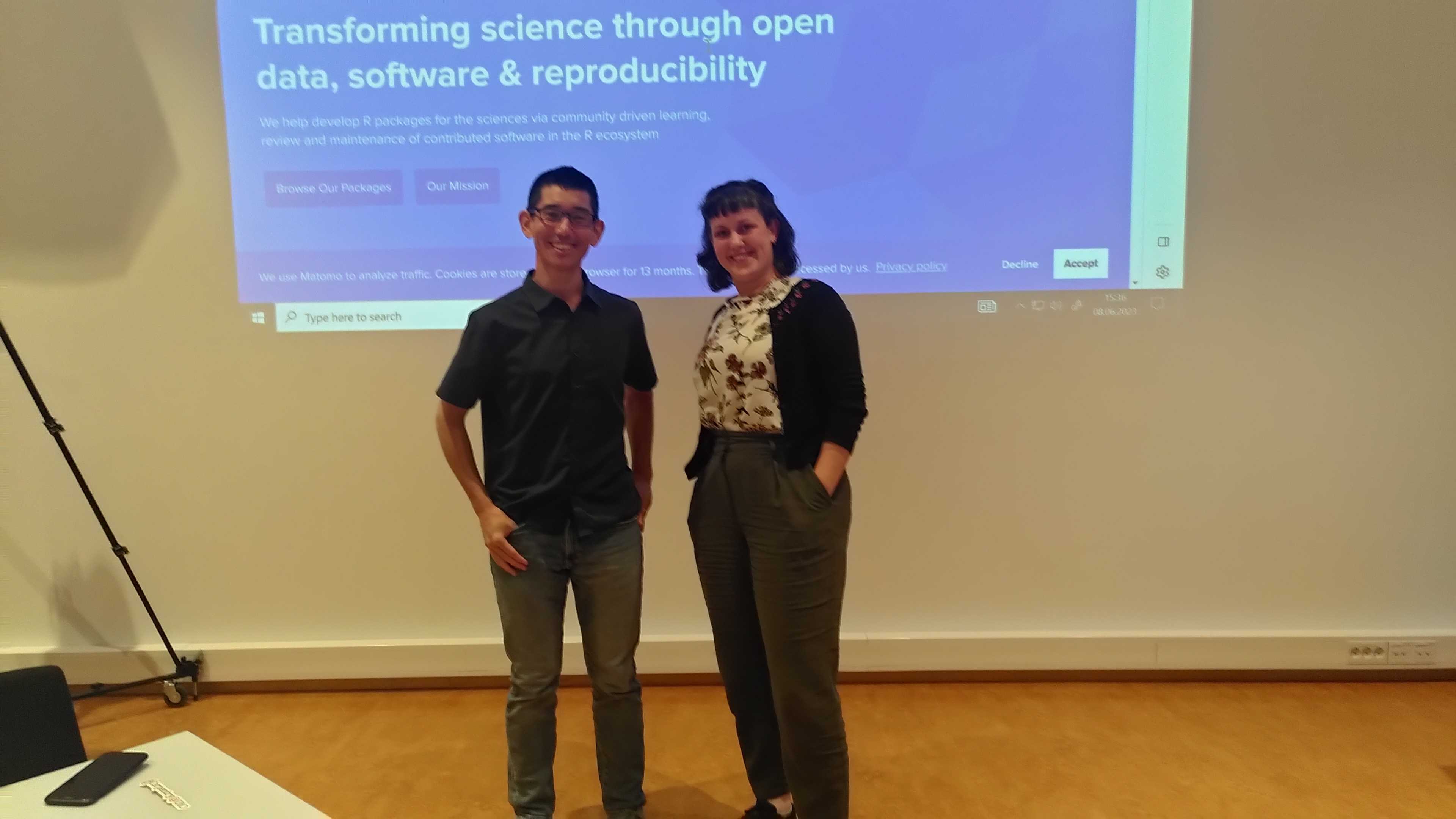 Joel and Mo posing with grins after the workshop in front of a large projector screen showing the rOpenSci website behind them.